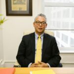 Middle-aged man in business attire sitting at an office desk with a blurred background.